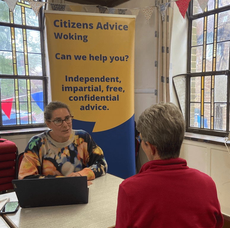Two people sit opposite each other at a table. One person, with a laptop, appears to be giving advice. Behind them is a banner for Citizens Advice Woking offering independent, impartial, free, confidential advice.